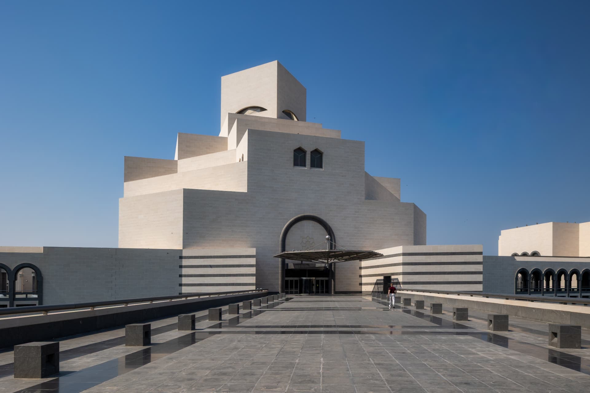 Stepped limestone facade and arched entrance of the Museum of Islamic Art in Doha under a clear blue sky, framed by a stone promenade