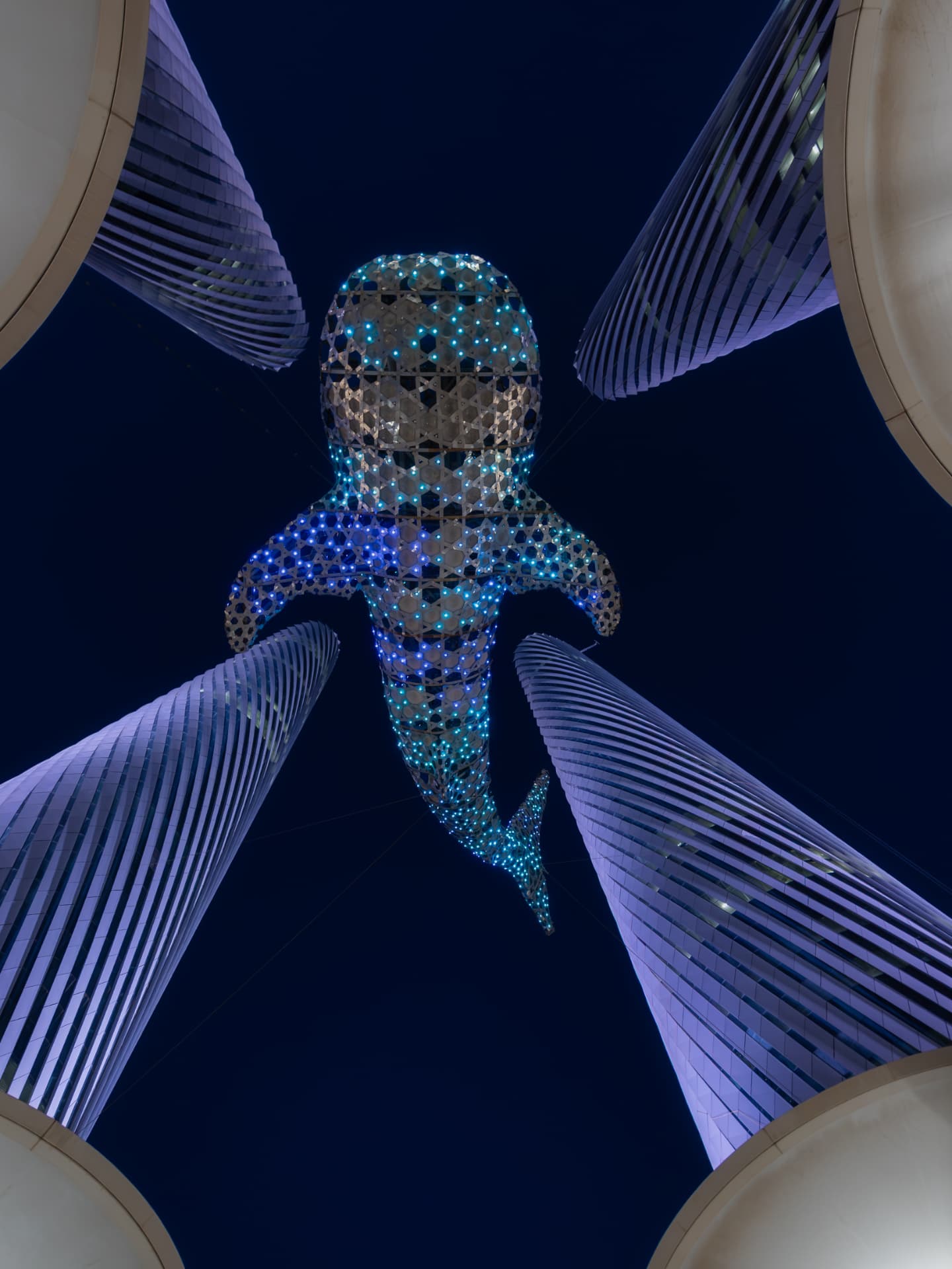 Looking up at an illuminated whale shark lattice sculpture suspended between four spiralling towers lit violet against the night sky
