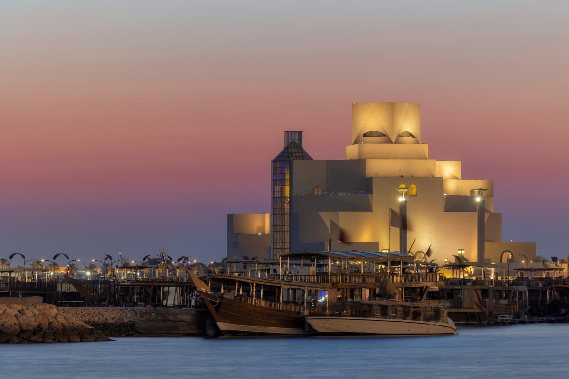 Museum of Islamic Art glowing gold at dusk under a pink sky, with traditional wooden dhows moored in Doha harbour