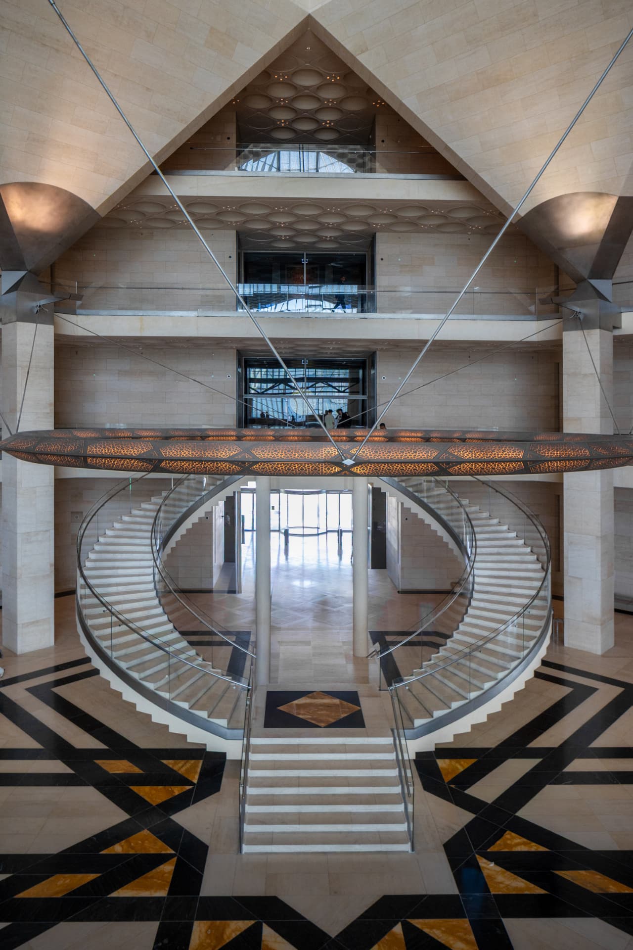 Elevated view of the Museum of Islamic Art's double spiral staircase beneath a glowing geometric chandelier and patterned marble floor
