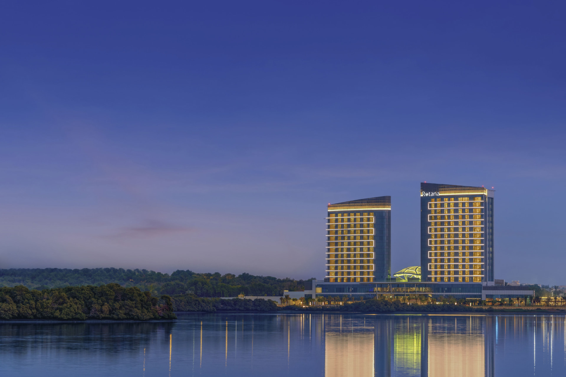 Rotana Ras Al Khaimah, The Mangroves after the refresh — the two illuminated towers at blue hour above the mangrove fringe, reflected in the still water of the channel