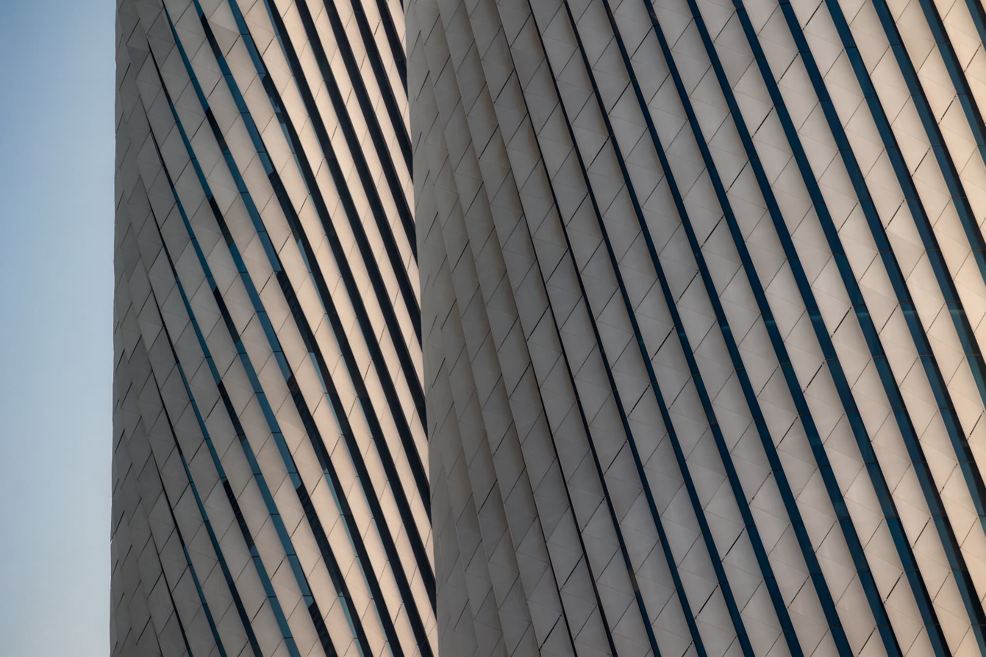 Close-up of a twisting tower facade with diagonal metal fins and dark glazing catching warm evening light against a pale sky