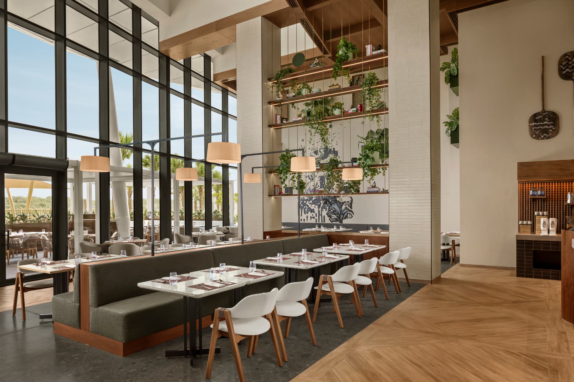 Terra restaurant at Rotana Ras Al Khaimah after the refresh — double-height glazing onto palms and mangroves balanced against interior ambient light, planted shelving, pendant lamps and set dining tables
